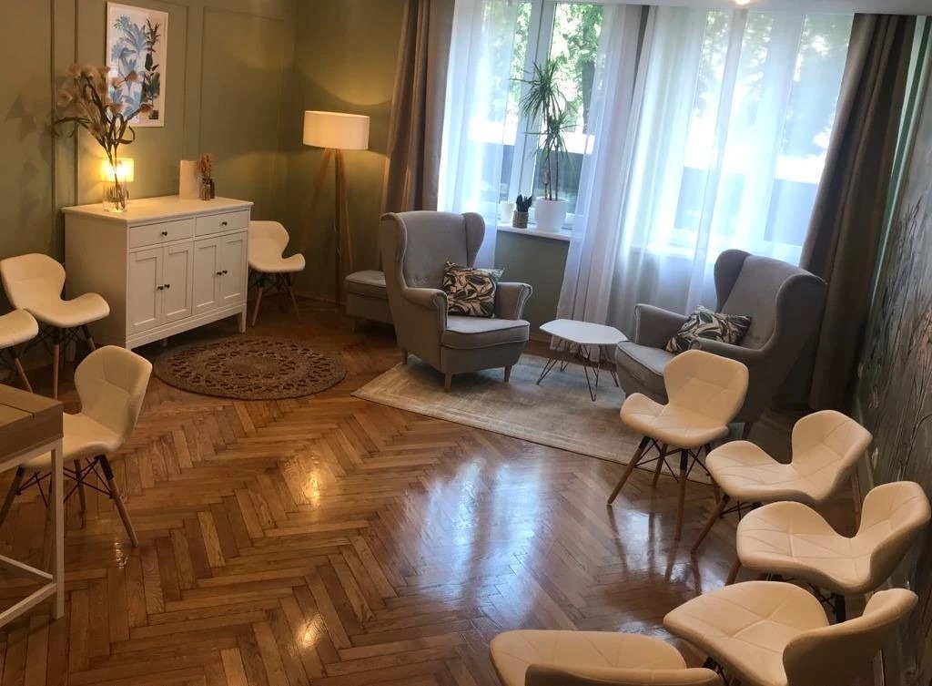 Jasne wnętrze gabinetu terapeutycznego z drewnianą podłogą w jodełkę, krzesłami w stylu skandynawskim i dwoma fotelami uszakami, oświetlone dziennym światłem wpadającym przez okno.