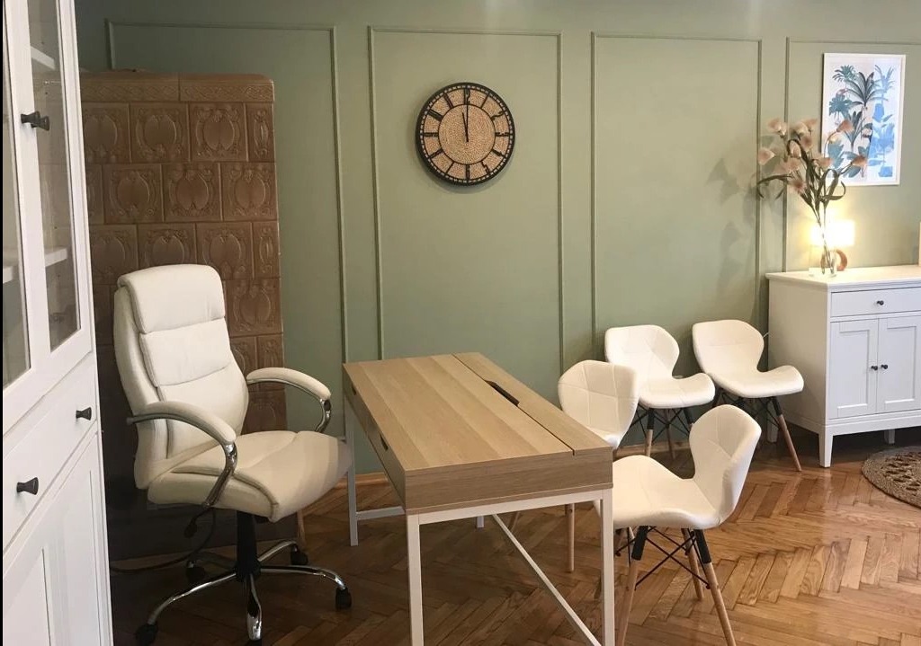 Jasne wnętrze gabinetu z biurkiem, krzesłami i ozdobnym piecem kaflowym w tle.