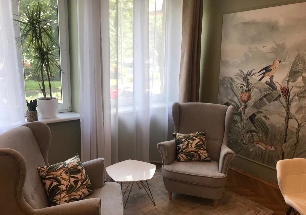 Jasny pokój z dwoma fotelami typu uszak, stolikiem kawowym o trójkątnym blacie i tropikalną tapetą w tle. W oknie widoczna zieleń.