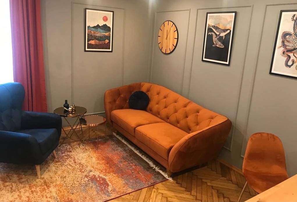 Pomarańczowa sofa z weluru z pikowaniem w gabinecie z szarymi ścianami, drewnianym zegarem i obrazami w czarnych ramach.