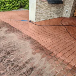 Częściowo umyta, czerwona kostka brukowa przed budynkiem z użyciem myjki ciśnieniowej. Widoczny efekt czyszczenia.