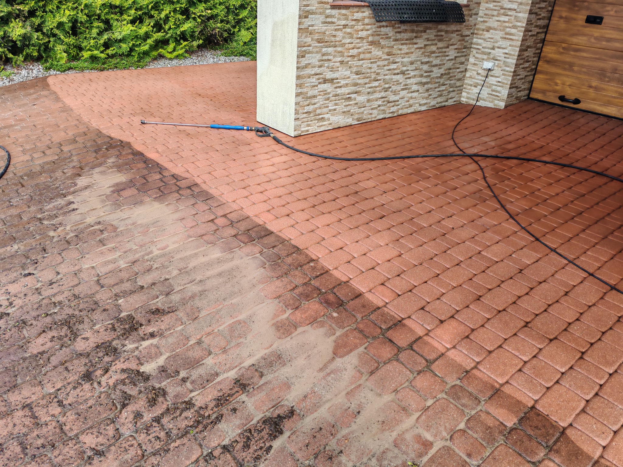 Częściowo umyta, czerwona kostka brukowa przed budynkiem z użyciem myjki ciśnieniowej. Widoczny efekt czyszczenia.