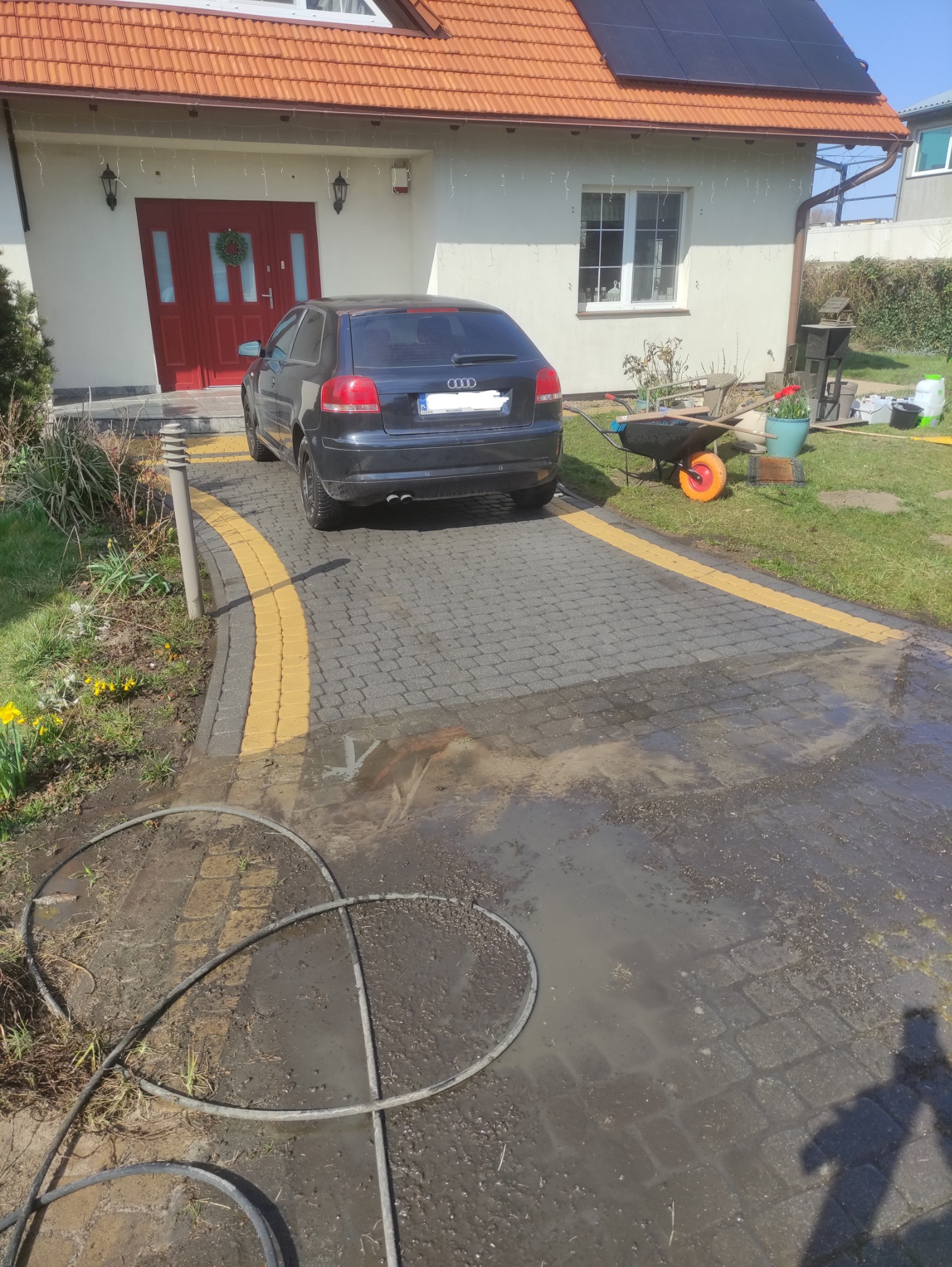Umyta część kostki brukowej przed domem z ciemnym autem, na pozostałej części widoczne zabrudzenia i woda z węża ogrodowego.