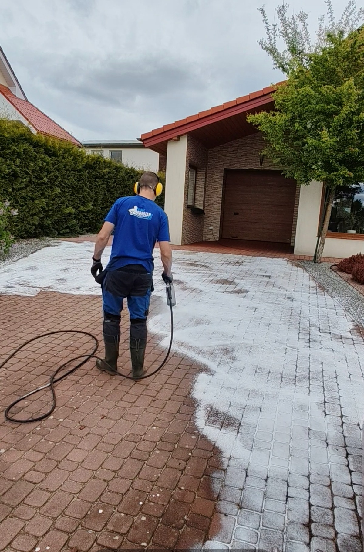 Pracownik w słuchawkach czyści kostkę brukową myjką ciśnieniową. Widoczny efekt czyszczenia: część kostki jest już umyta, a część jeszcze brudna. Dom z garażem w tle.