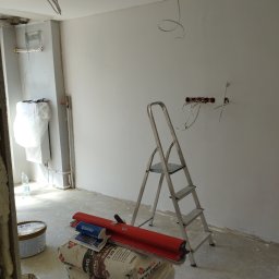 Krzysztof Martowicz - Wnętrze w trakcie szpachlowania: worki z zaprawą, drabina, narzędzia, widoczne kable elektryczne w ścianie. Ściany przygotowane do malowania.