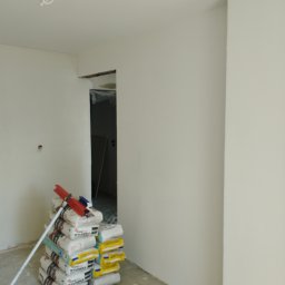 Krzysztof Martowicz - Wnętrze po szpachlowaniu i malowaniu na biało, widoczny stos worków z zaprawą, wiadro z resztkami farby i mop oparty o ścianę. Widok na otwór drzwiowy.