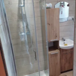 Nowoczesna kabina prysznicowa z deszczownicą, umywalka z szafką w łazience wykończonej płytkami imitującymi drewno. Widok na lustro i akcesoria łazienkowe.