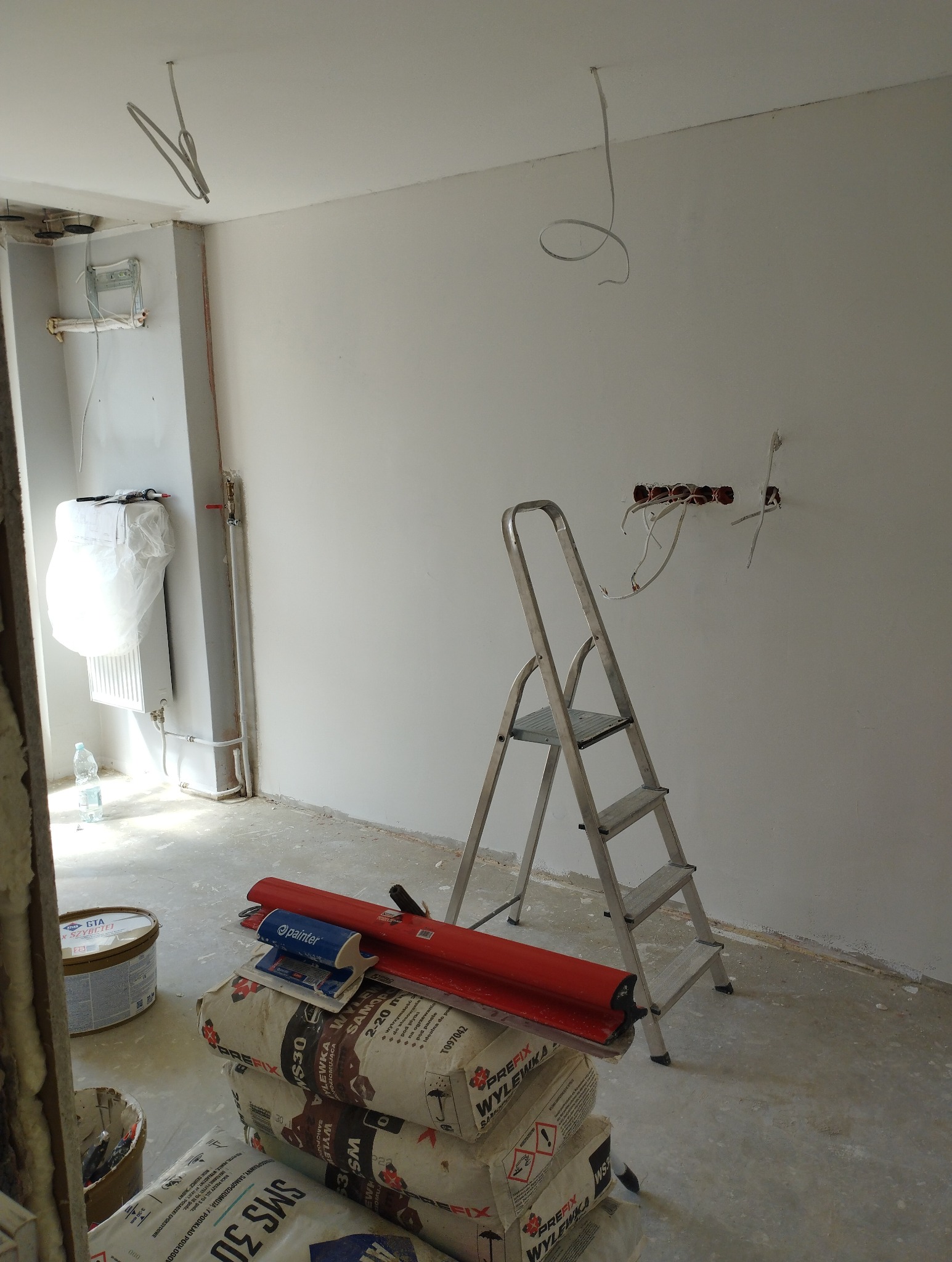 Wnętrze w trakcie szpachlowania: worki z zaprawą, drabina, narzędzia, widoczne kable elektryczne w ścianie. Ściany przygotowane do malowania.