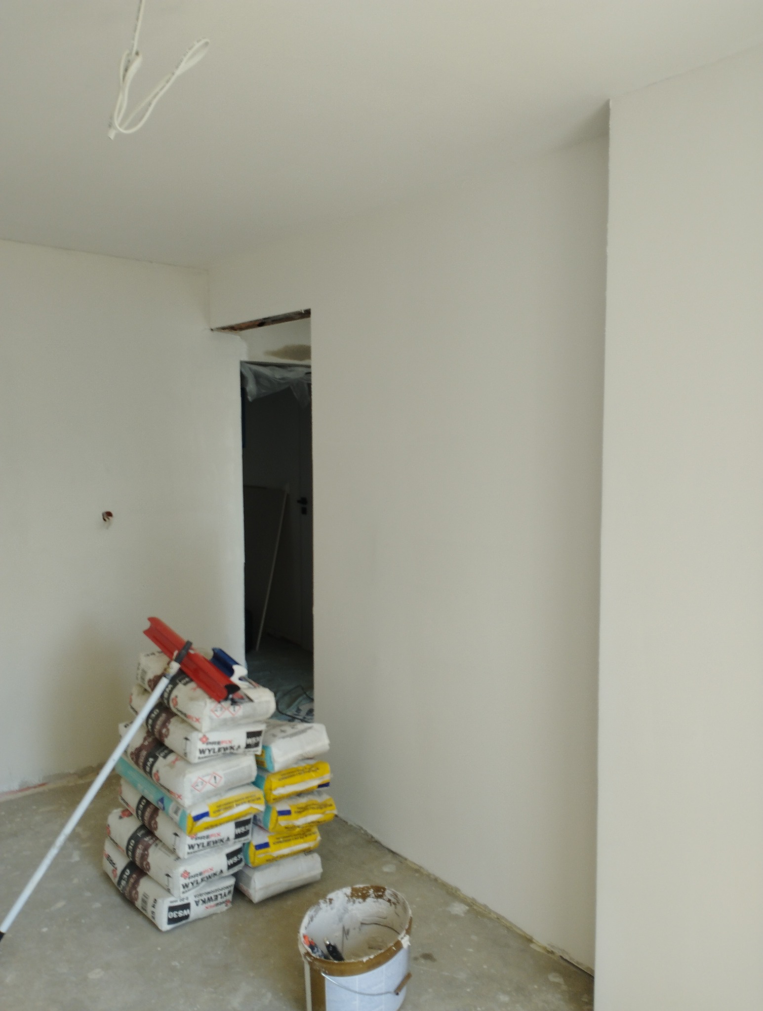 Wnętrze po szpachlowaniu i malowaniu na biało, widoczny stos worków z zaprawą, wiadro z resztkami farby i mop oparty o ścianę. Widok na otwór drzwiowy.