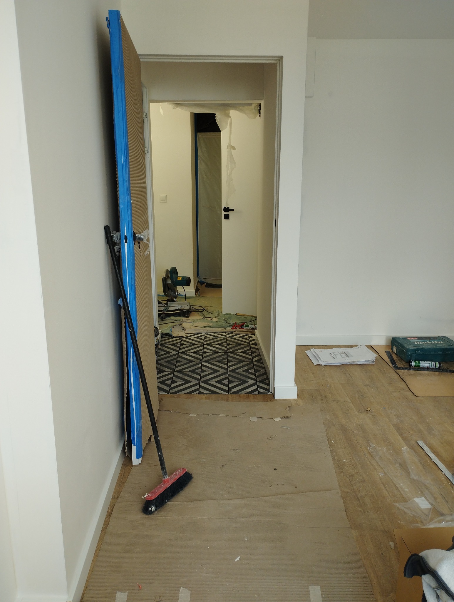 Wnętrze w trakcie remontu: zabezpieczona podłoga, ościeżnica z niebieską taśmą, widoczna nowa podłoga we wzory i narzędzia w tle. Etap prac wykończeniowych.
