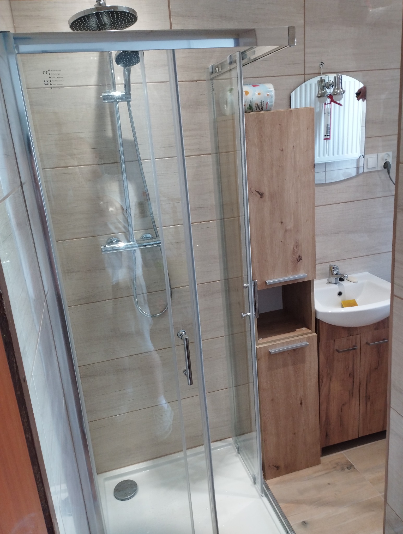 Nowoczesna kabina prysznicowa z deszczownicą, umywalka z szafką w łazience wykończonej płytkami imitującymi drewno. Widok na lustro i akcesoria łazienkowe.