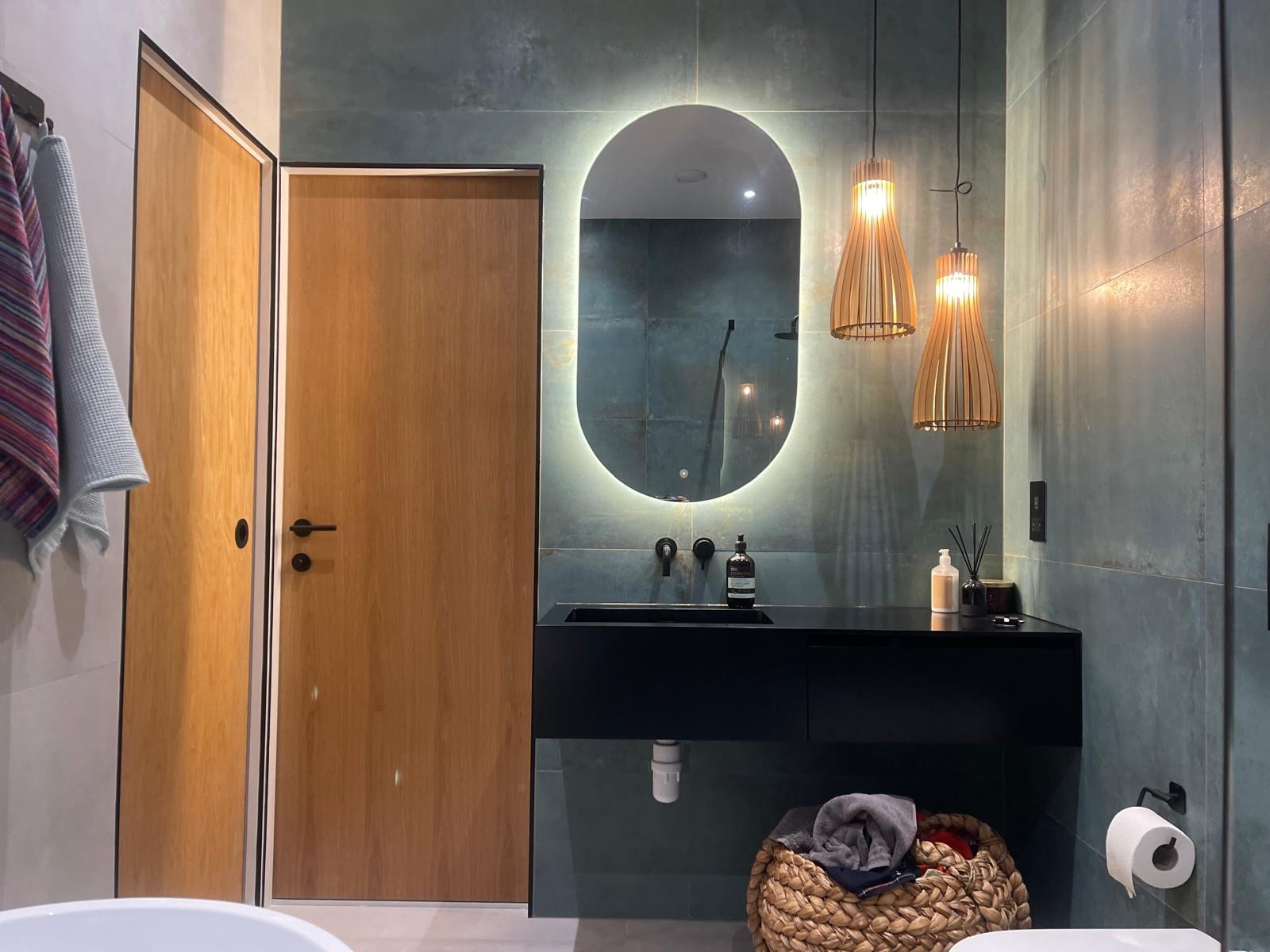 Nowoczesna łazienka z drewnianymi drzwiami, owalnym lustrem z podświetleniem LED i czarną szafką pod umywalką. Wiszące lampy z drewnianymi elementami.