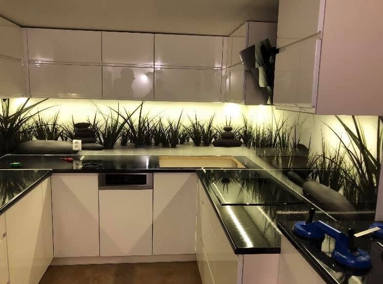 Nowoczesna kuchnia z czarnym blatem i szklanym panelem ściennym z motywem trawy, podświetlonym od góry. Widoczne narzędzia montażowe.