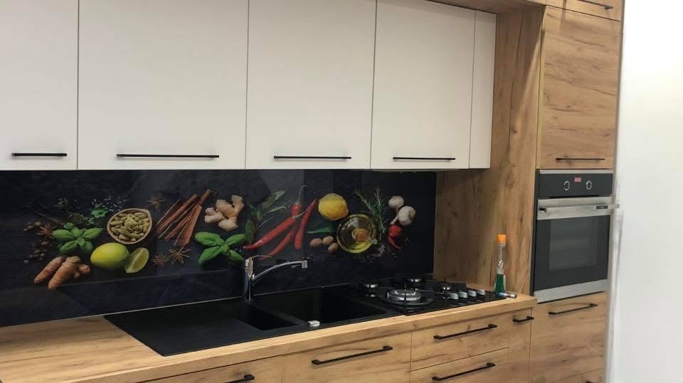 Kuchnia z drewnianymi szafkami i czarnym panelem szklanym nad blatem, zdobionym motywem przypraw i ziół. Nowoczesny design, czarne uchwyty, piekarnik w zabudowie.