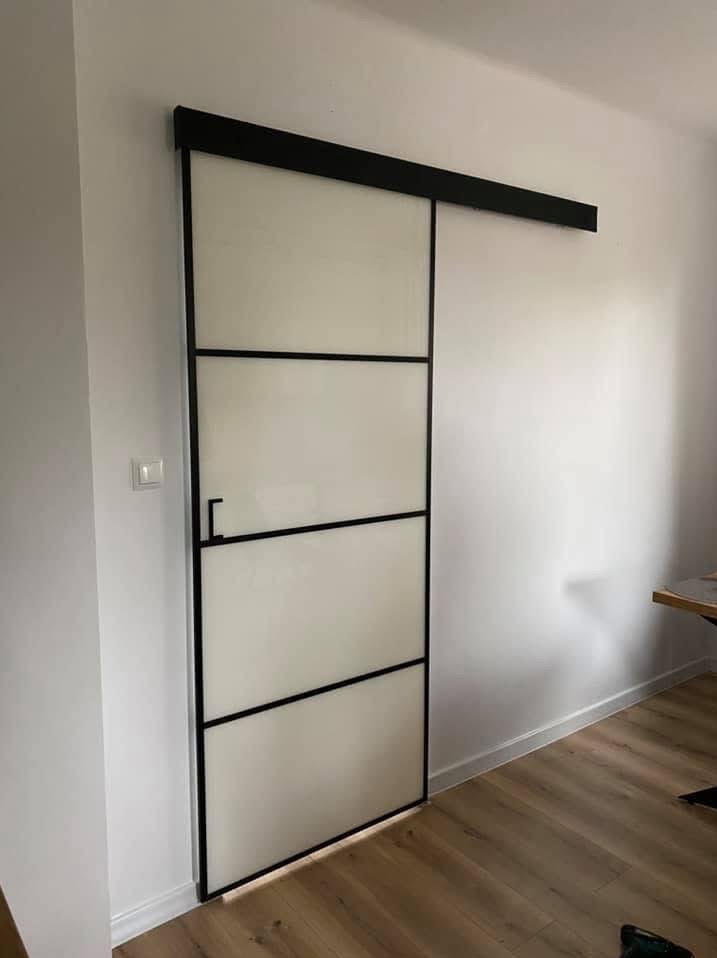 Nowoczesne, przesuwne drzwi z matowego szkła w czarnej ramie, zamontowane na szynie. Minimalistyczny design, jasne wnętrze, drewniana podłoga.