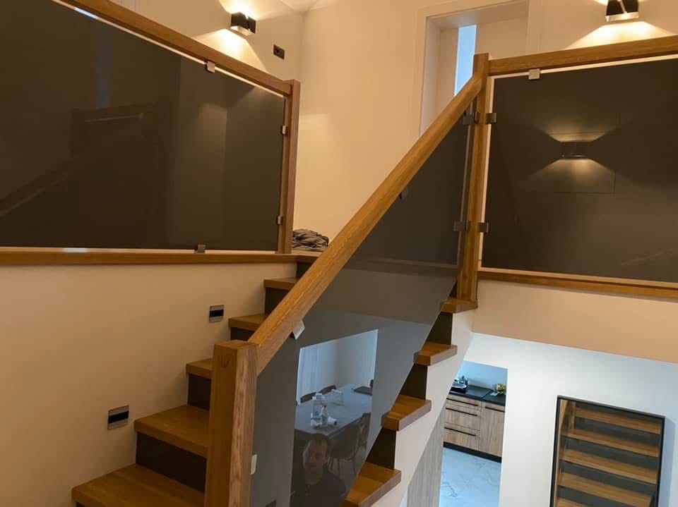 Nowoczesna balustrada szklana z drewnianymi elementami na schodach w jasnym wnętrzu. Widoczne oświetlenie punktowe i minimalistyczny design.