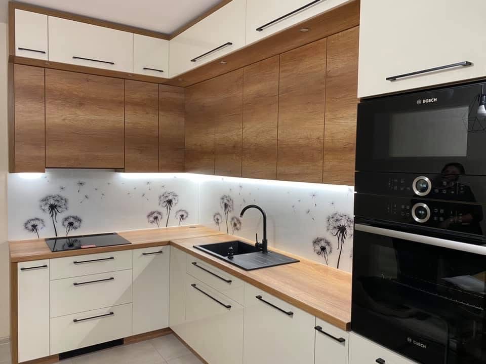 Nowoczesna kuchnia w bieli i drewnie z motywem dmuchawców na szklanym panelu ściennym, czarne akcenty, oświetlenie LED podszafkowe, zabudowa piekarnika Bosch.