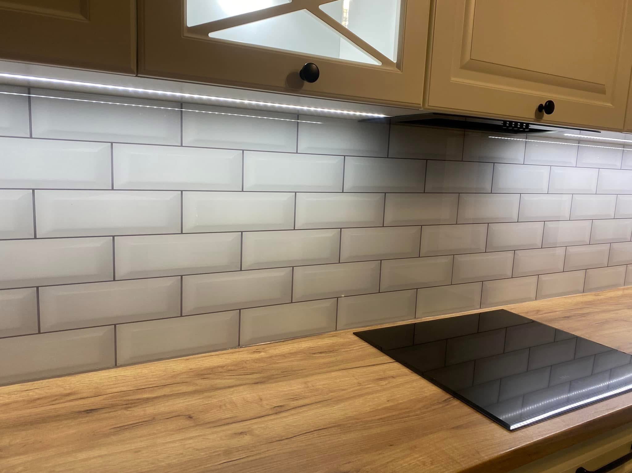 Białe, błyszczące płytki typu metro z ciemną fugą na ścianie nad drewnianym blatem kuchennym z czarną płytą indukcyjną. Pod szafkami ledowe oświetlenie.