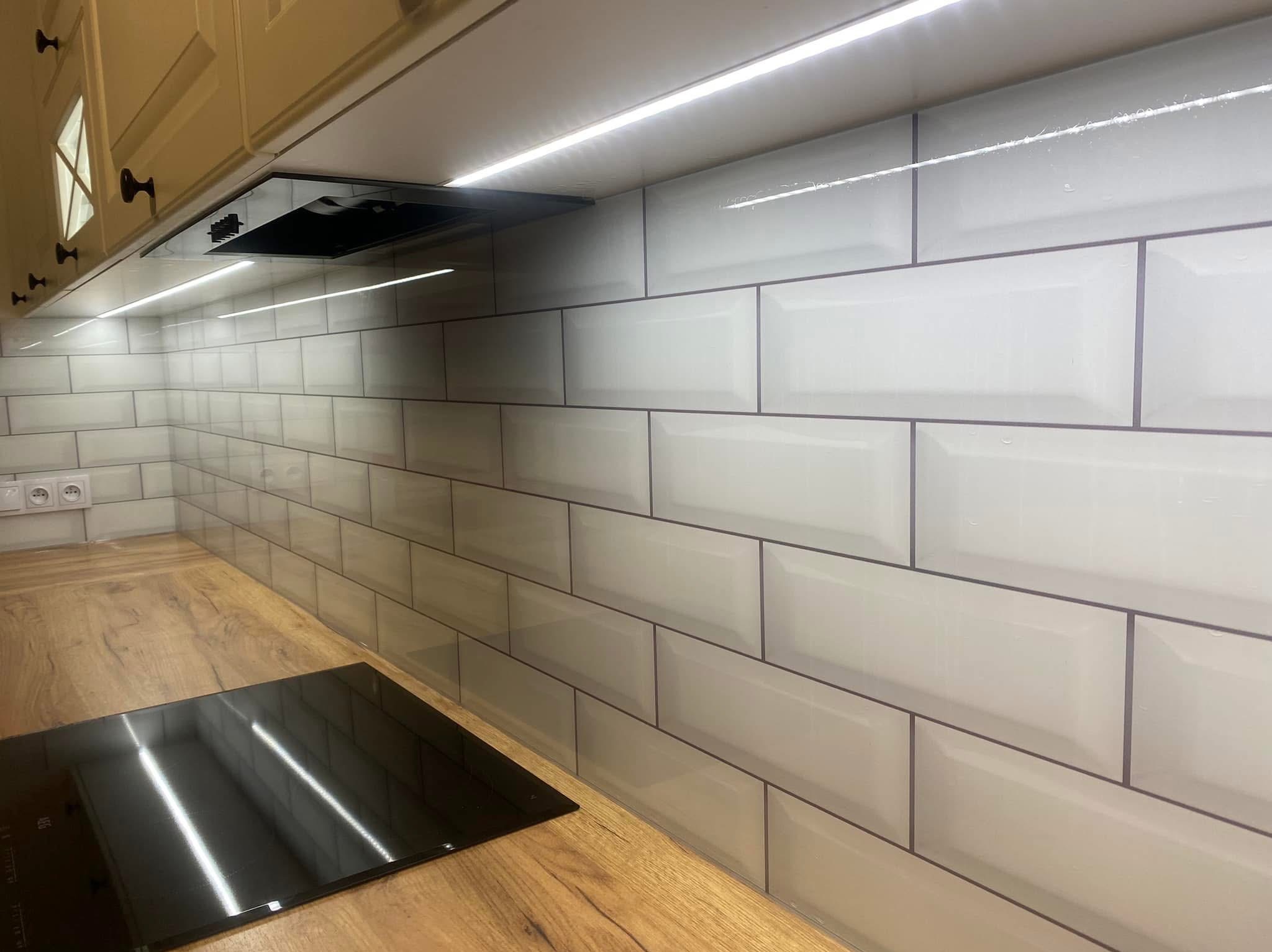 Białe, prostokątne płytki na ścianie w kuchni, ułożone w cegiełkę, z drewnianym blatem i czarną płytą indukcyjną. Oświetlenie LED pod szafkami.