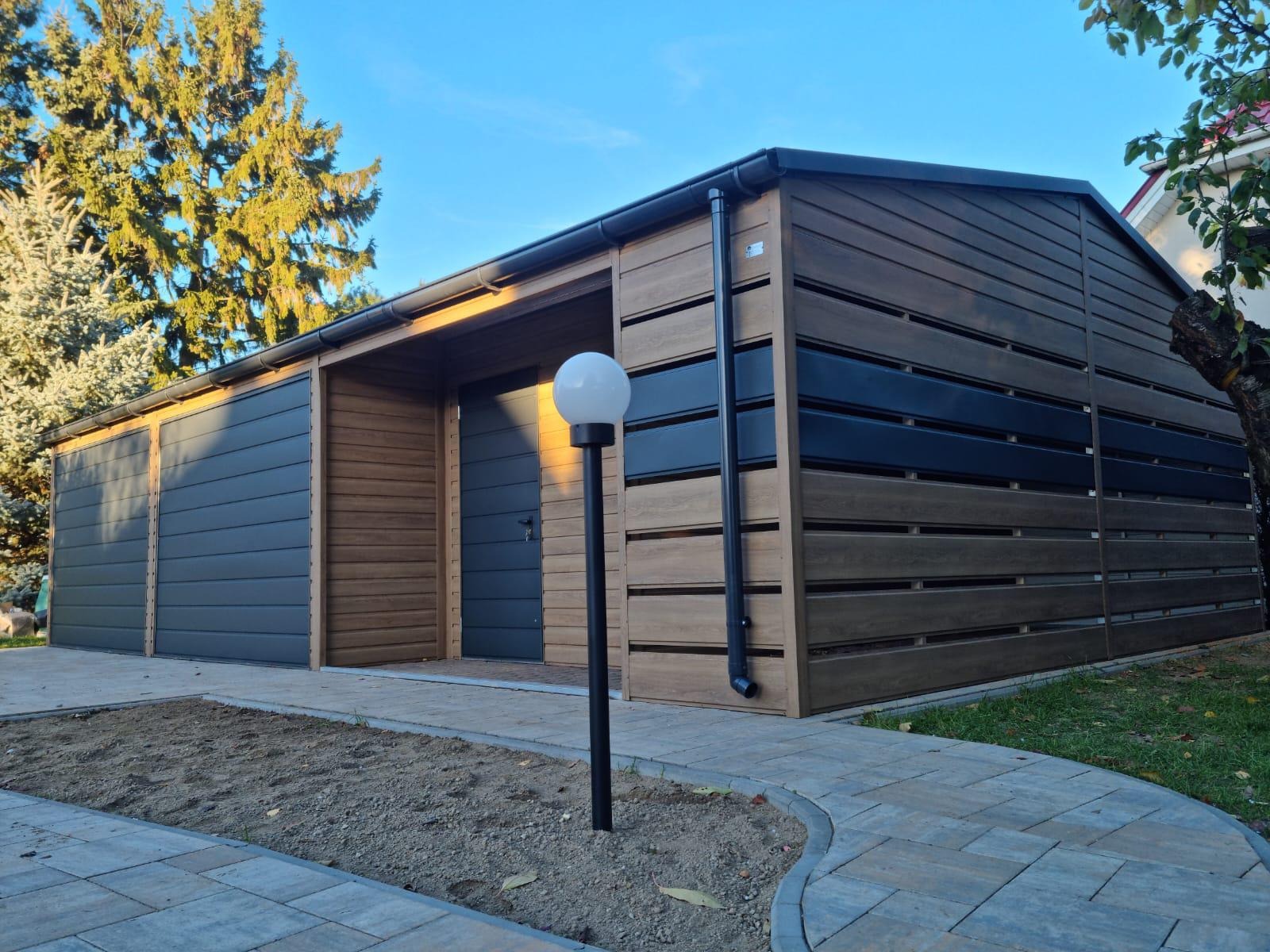 Nowoczesny garaż z blachy drewnopodobnej, z dwoma bramami segmentowymi i drzwiami wejściowymi, oświetlony lampą kulą, z widoczną kostką brukową i elementami zieleni.