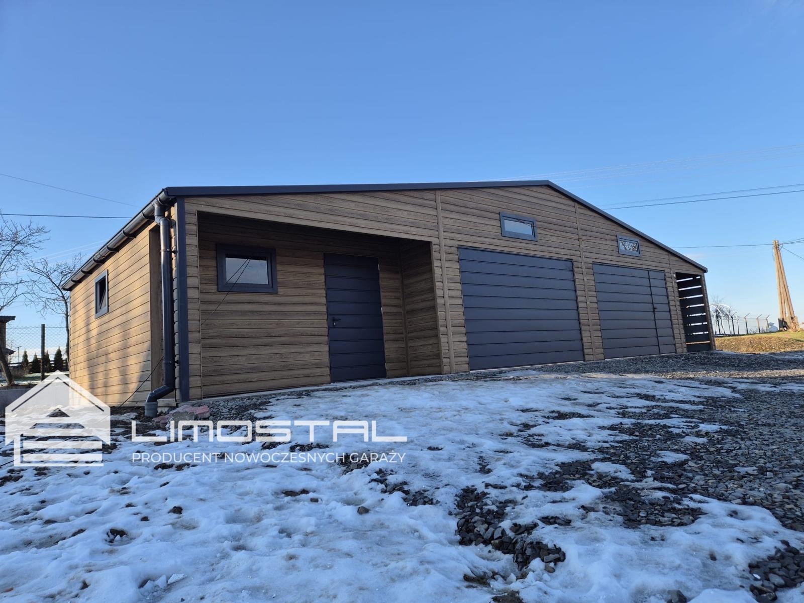Nowoczesny garaż dwustanowiskowy z drewnopodobną elewacją i dwiema bramami segmentowymi w kolorze antracytowym, widoczny częściowo ośnieżony teren i logo producenta.