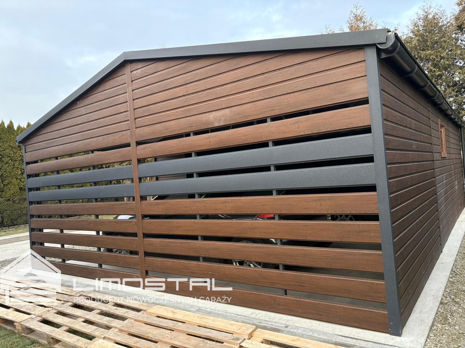 Nowoczesny garaż blaszany z poziomymi panelami imitującymi drewno, szara konstrukcja ramy, widoczny fragment samochodu wewnątrz, paleta drewniana na pierwszym planie z logo producenta.