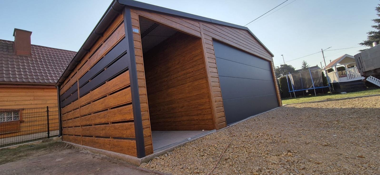 Nowoczesny garaż z drewnopodobną elewacją i segmentową bramą w kolorze antracytowym, otoczony żwirowym podłożem, z widokiem na dom i trampolinę w tle.