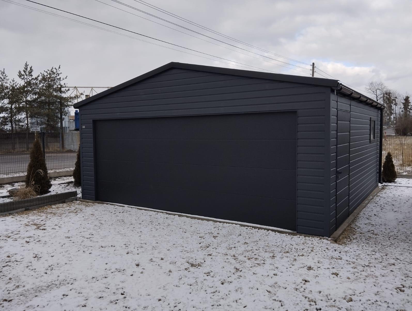 Gotowy, ciemnoszary garaż z blachy trapezowej z bramą segmentową, widoczny w zimowym krajobrazie z resztkami śniegu na ziemi.