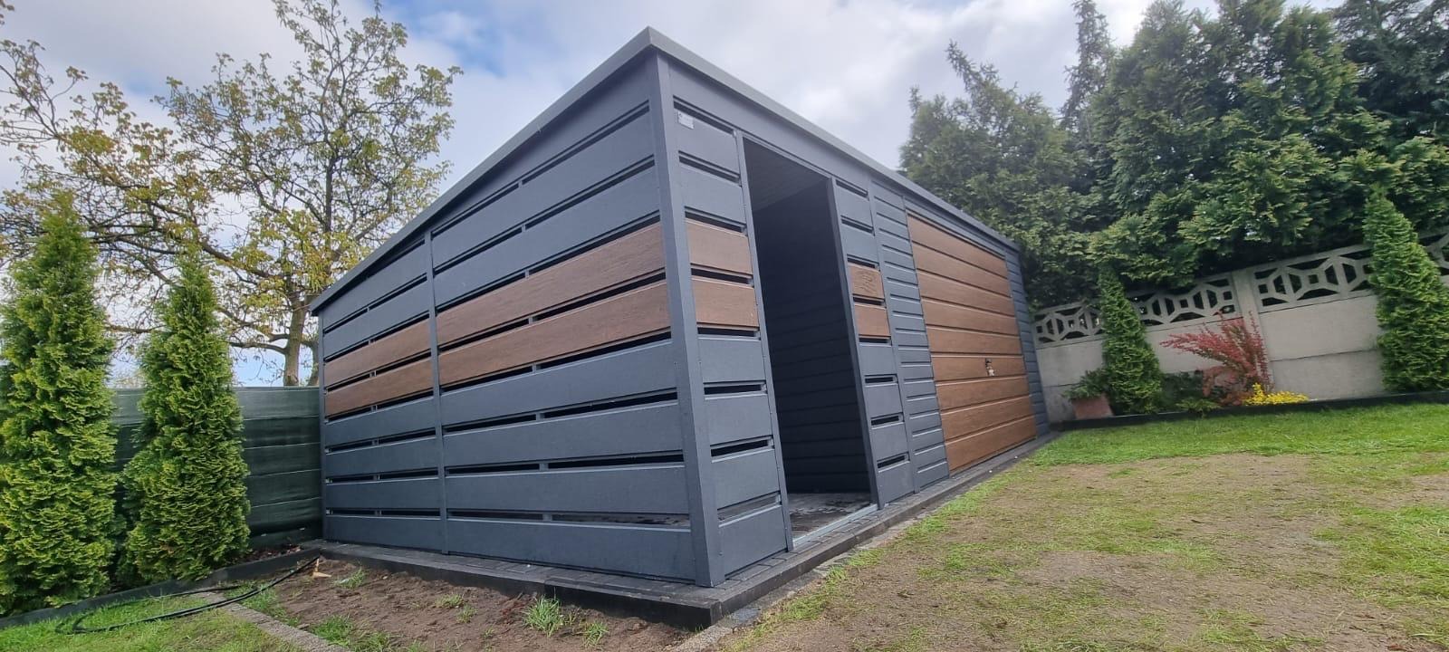 Nowoczesna konstrukcja z szarych i brązowych paneli, przypominająca domek narzędziowy lub mały garaż, z otwartymi drzwiami i widocznym trawnikiem oraz drzewami w tle.