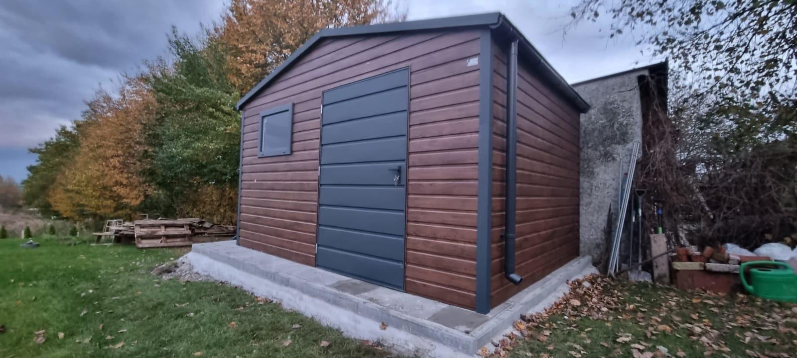 Gotowy, brązowy budynek gospodarczy z szarą, segmentową bramą i oknem, na betonowej wylewce, z rynnami, na tle jesiennego ogrodu.