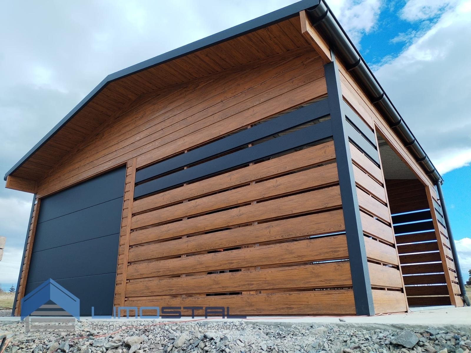 Nowoczesny garaż blaszany z drewnopodobną elewacją, czarną bramą segmentową i otwartą przestrzenią z boku, widoczny fragment logo producenta na dole zdjęcia.