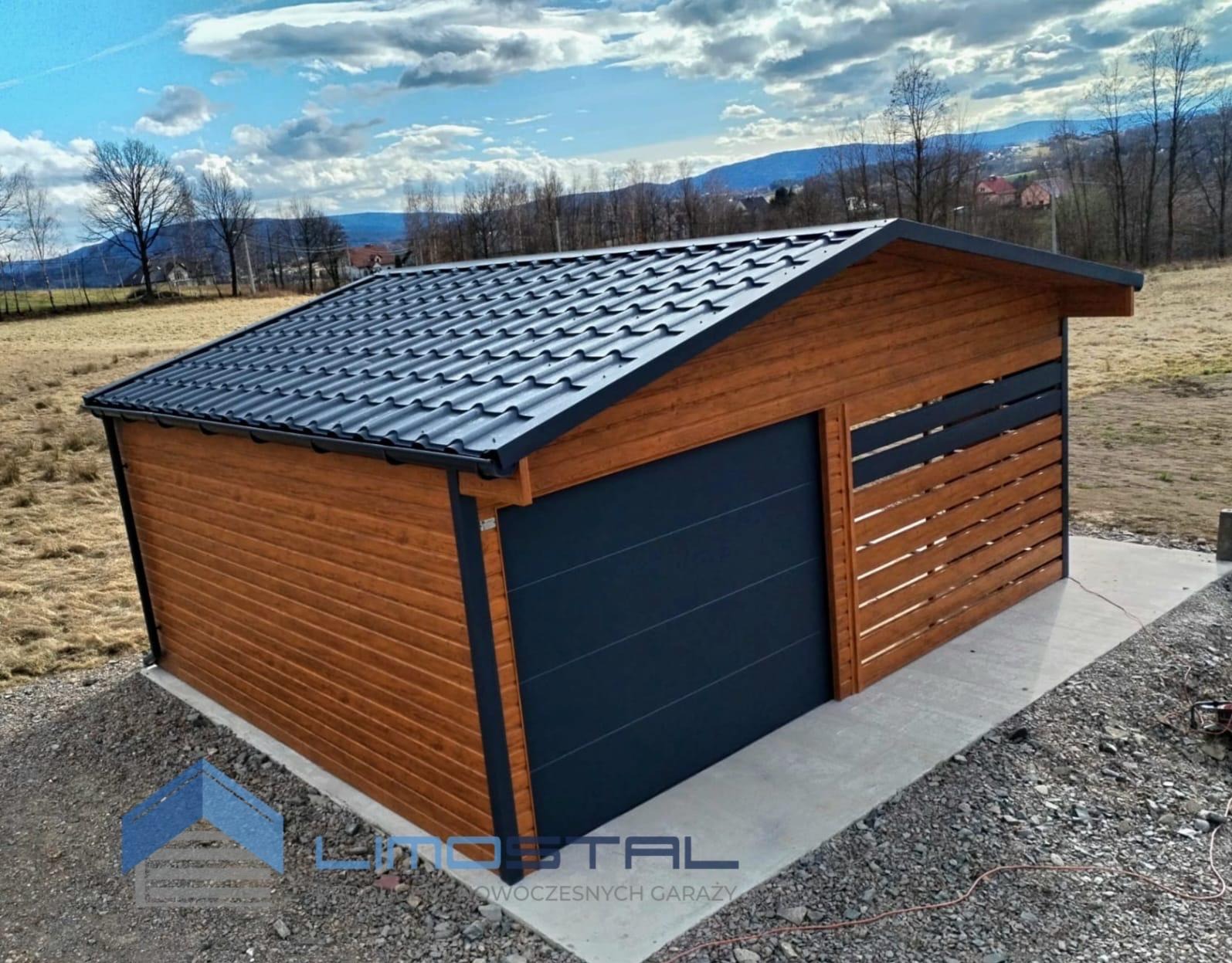 Drewniany garaż z ciemnoszarą bramą segmentową i dachem z blachy trapezowej, widok z boku na betonowej wylewce, w tle krajobraz z górami.