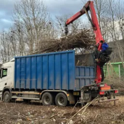 Ciężarówka z niebieską skrzynią ładunkową, napełniana gałęziami za pomocą czerwonego dźwigu z operatorem w niebieskiej bluzie; w tle drzewa bez liści i zielony ekran akustyczny.