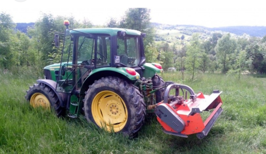 Zielony traktor z żółtymi felgami i pomarańczowym osprzętem stoi na łące z wysoką trawą, w tle zalesione wzgórza.