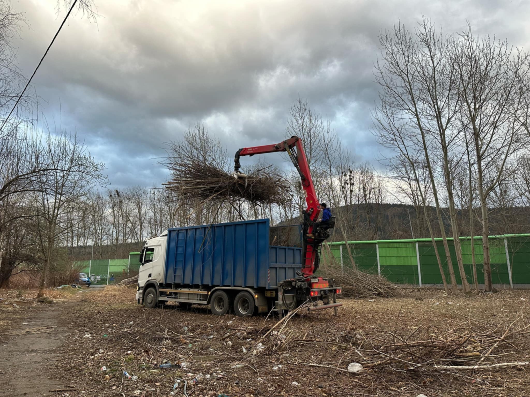Ciężarówka z kontenerem i dźwigiem załadowuje gałęzie po wycince drzew na tle pochmurnego nieba i zielonego ekranu akustycznego.
