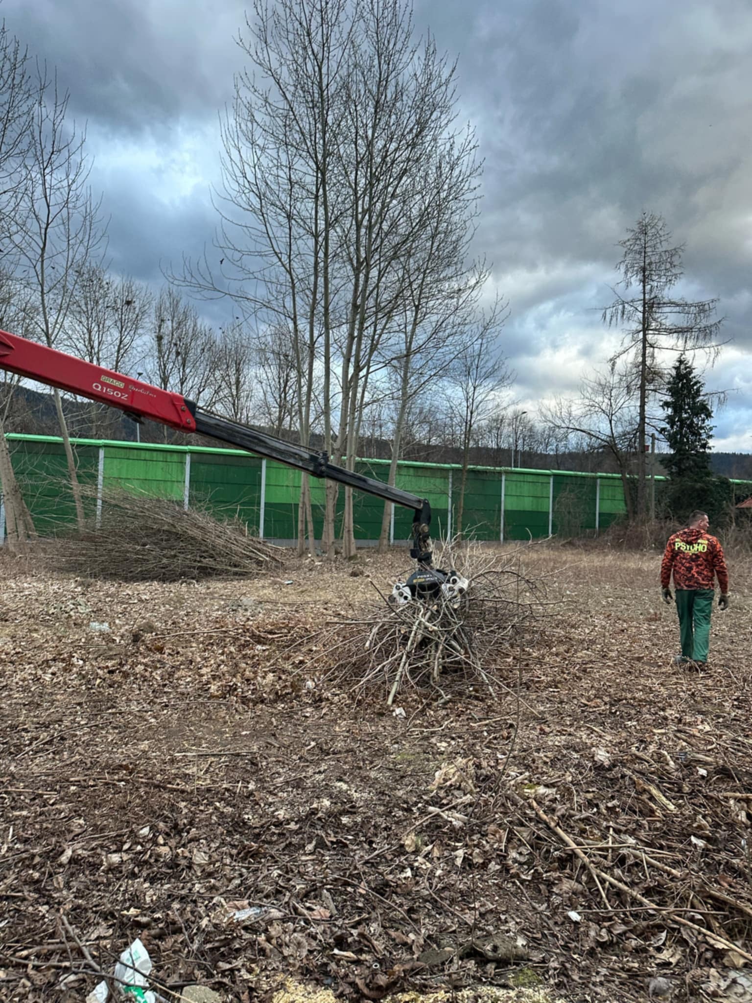 Czerwony dźwig chwyta gałęzie na zalesionym terenie, pracownik w zielonym kombinezonie oddala się od maszyny, w tle zielony ekran akustyczny i pochmurne niebo.