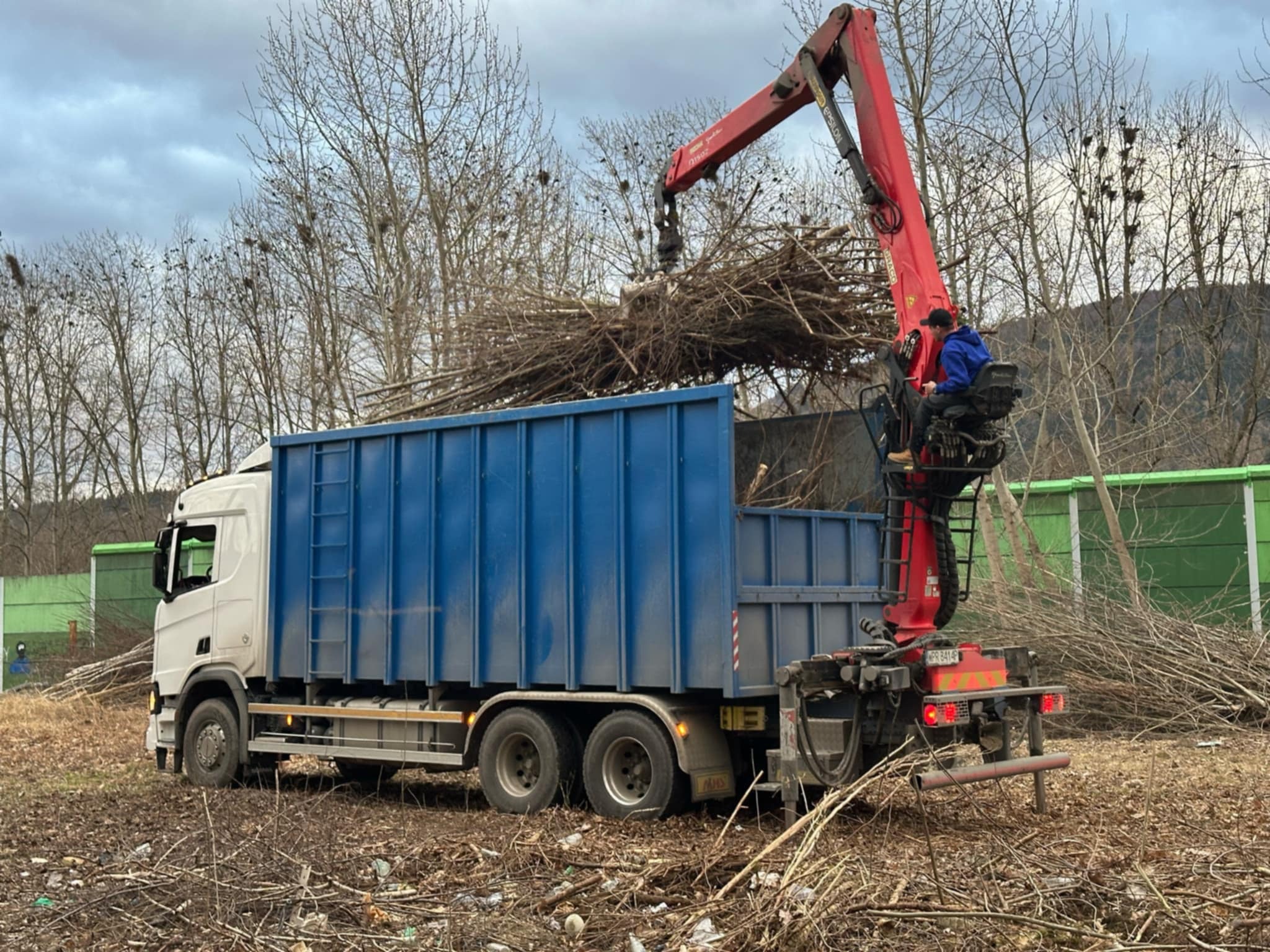 Ciężarówka z niebieską skrzynią ładunkową, napełniana gałęziami za pomocą czerwonego dźwigu z operatorem w niebieskiej bluzie; w tle drzewa bez liści i zielony ekran akustyczny.