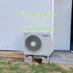 Zewnętrzna jednostka klimatyzacji Samsung na betonowych podstawach przy elewacji budynku. Widoczne elementy instalacji i trawa.