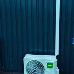 ECO Vento Dariusz Ogniewski - Klimatyzator Kaisai zamontowany na zewnątrz budynku z ciemną, metalową ścianą w tle, stojący na ceglanym podłożu. Widoczna rura odprowadzająca.