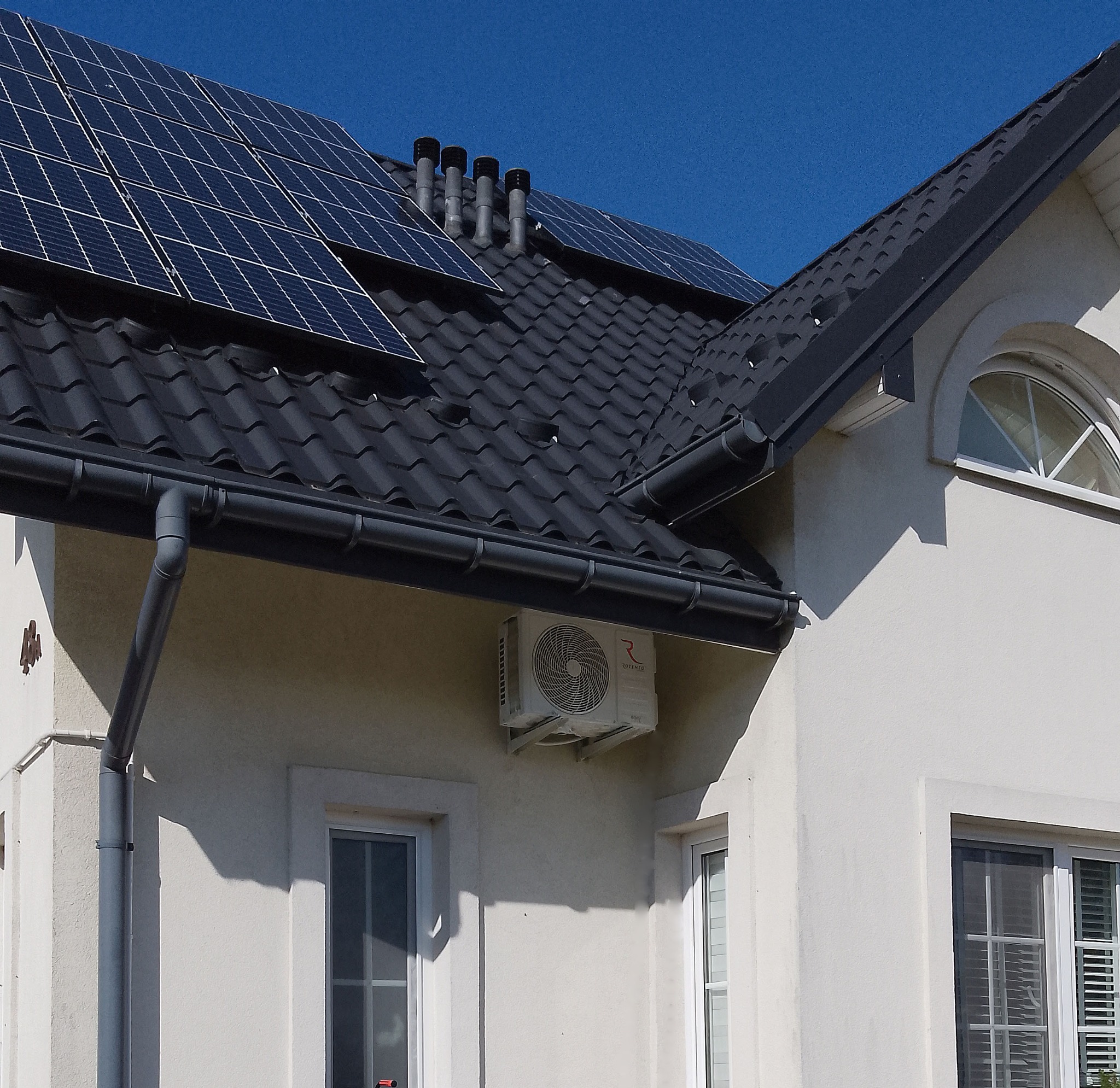 Biała ściana domu z zamontowaną jednostką zewnętrzną klimatyzacji Rotenso, widoczne okna, rynna spustowa i panele solarne na czarnym dachu.
