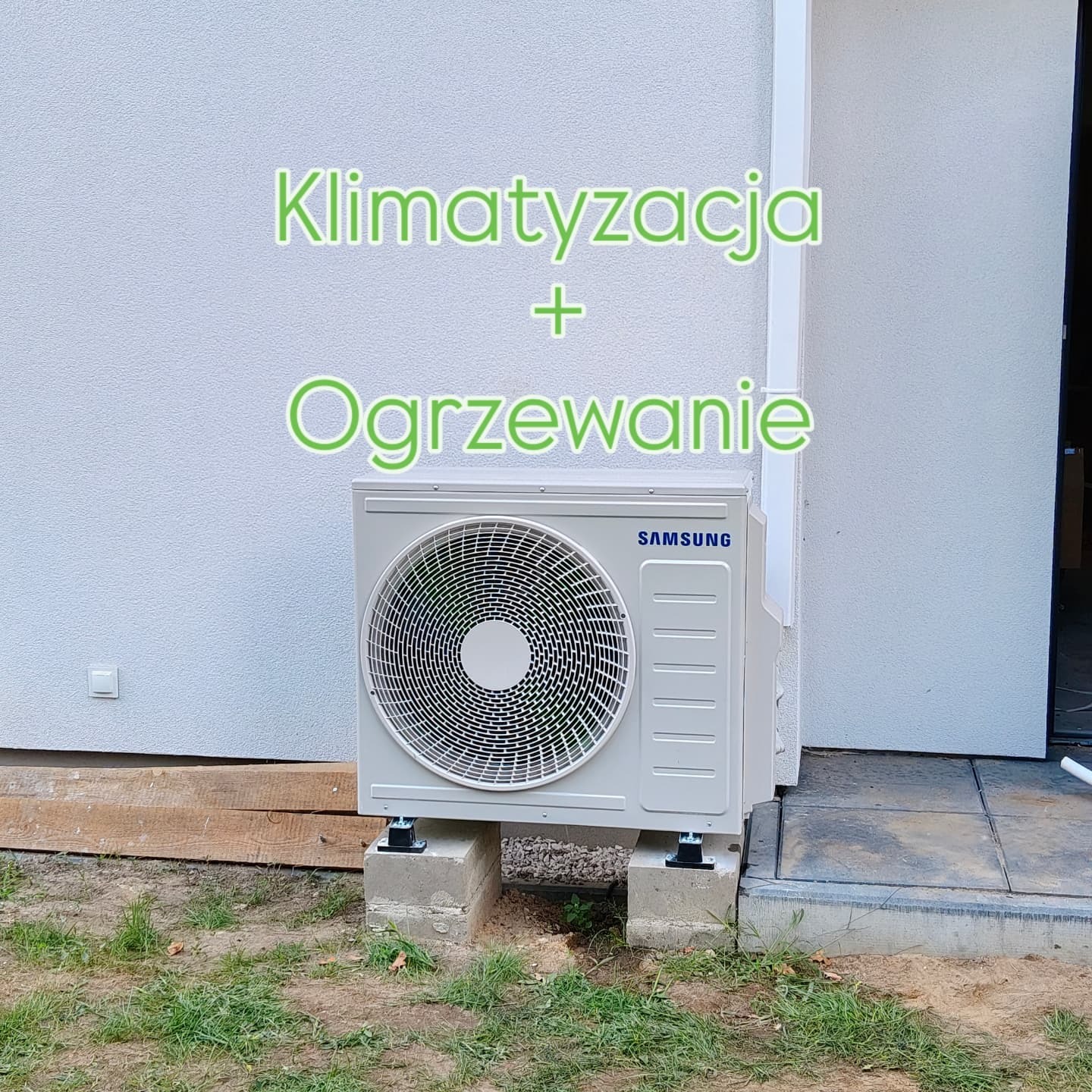 Zewnętrzna jednostka klimatyzacji Samsung na betonowych podstawach przy elewacji budynku. Widoczne elementy instalacji i trawa.