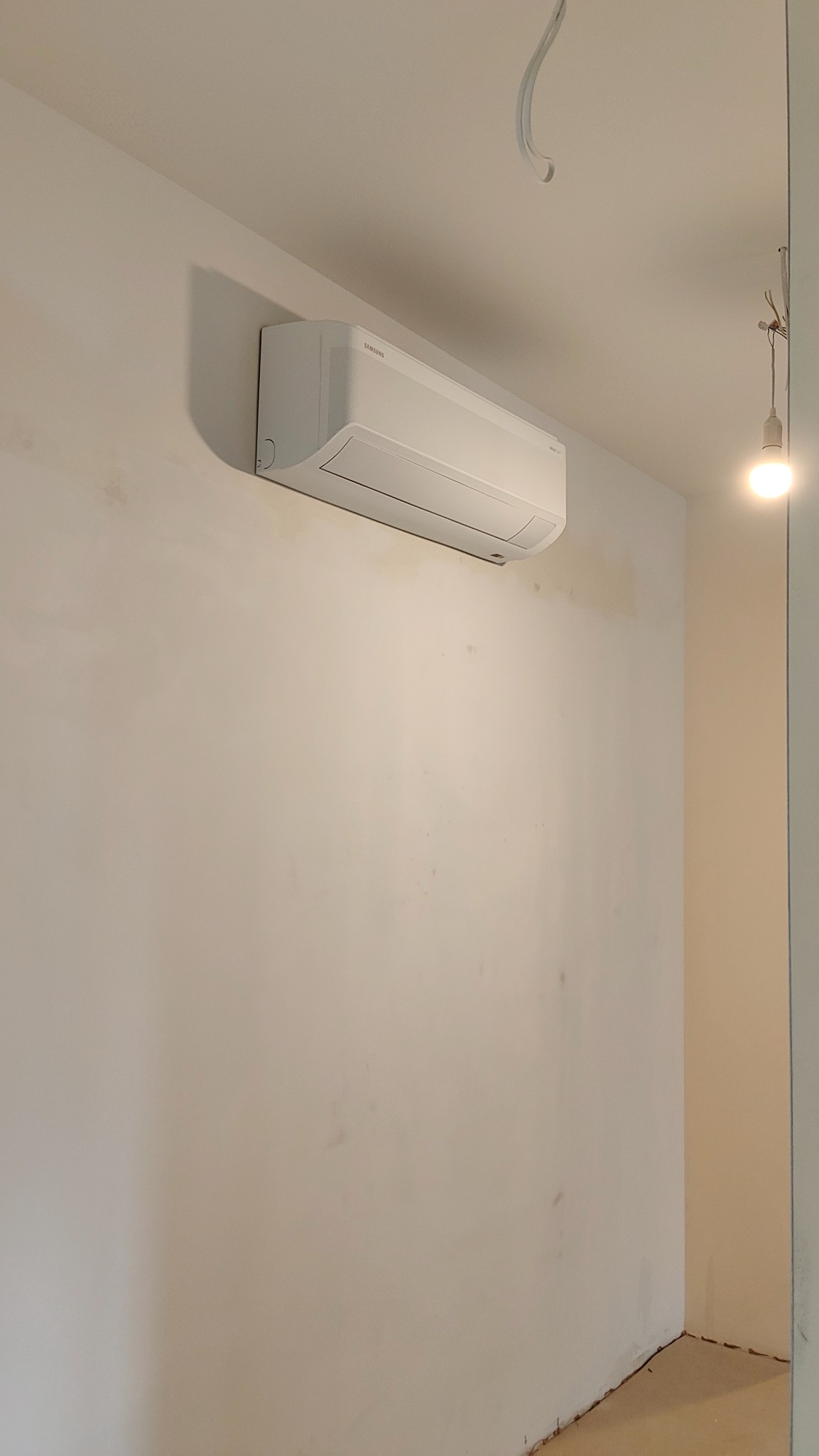 Biała klimatyzacja Samsung zamontowana na ścianie w trakcie remontu, widoczne ślady po pracach wykończeniowych. Minimalistyczny design, surowe wnętrze.