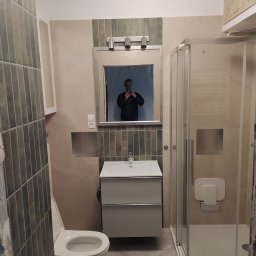 Szymon Kuprewicz - Wykończona łazienka z kabiną prysznicową, umywalką z szafką i toaletą. Widoczne szare płytki i lustro z odbiciem osoby robiącej zdjęcie.