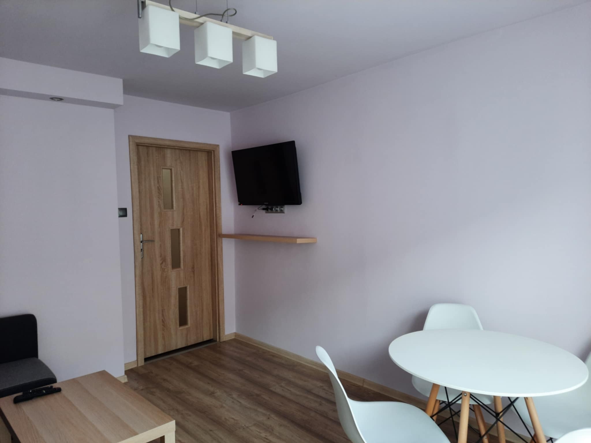 Jasne wnętrze pokoju z drewnianą podłogą, drzwiami z przeszkleniami, telewizorem na ścianie, białym okrągłym stołem i białymi krzesłami w stylu skandynawskim oraz lampą sufitową z trzema...