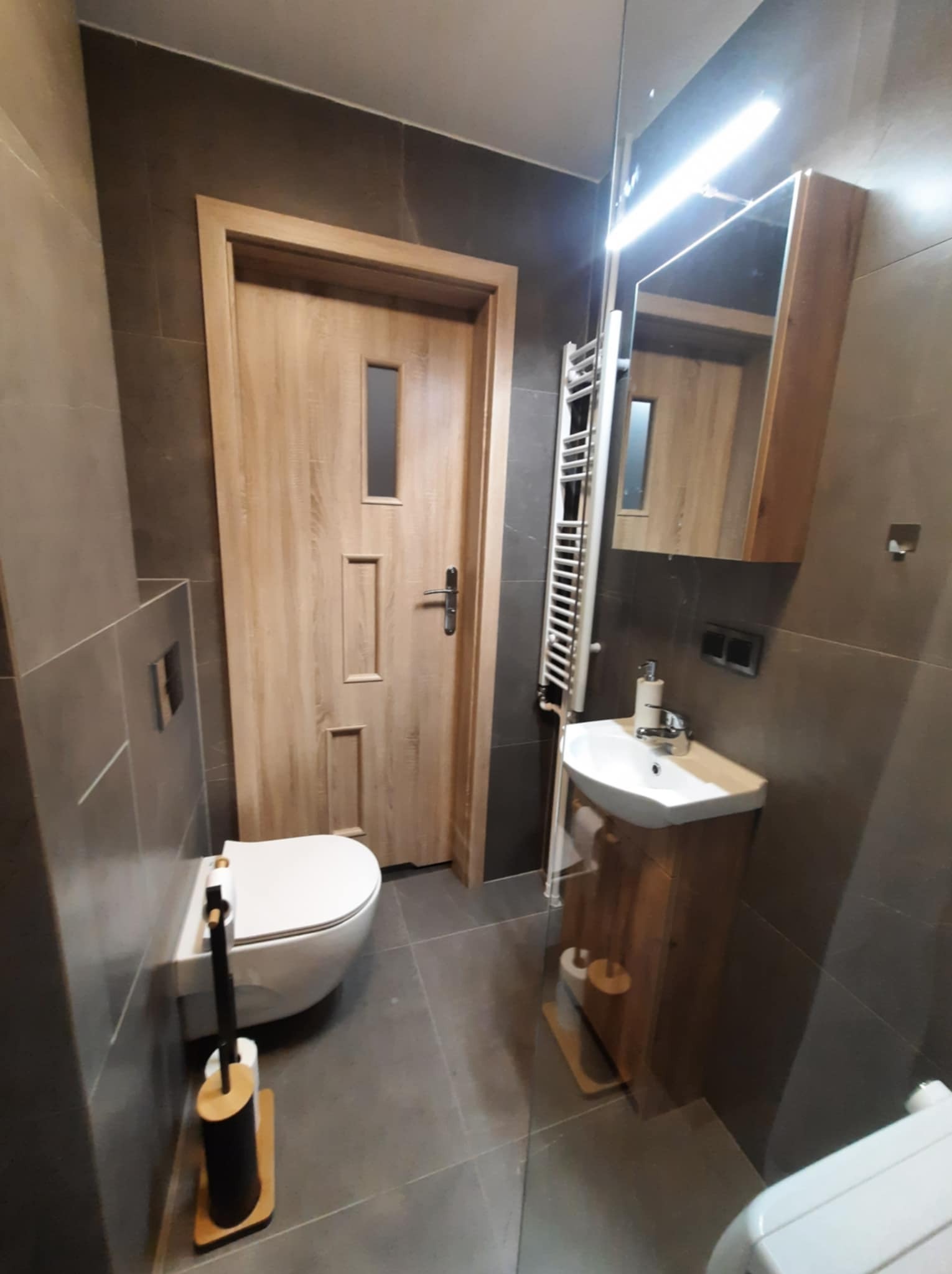 Nowoczesna łazienka z szarymi płytkami, drewnianymi akcentami na drzwiach i szafce pod umywalką, pionowy grzejnik drabinkowy i minimalistyczny zestaw toaletowy.