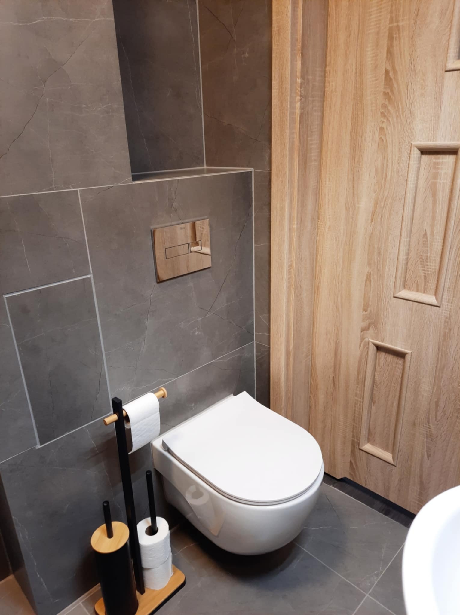 Nowoczesna łazienka z szarymi płytkami imitującymi marmur, podwieszana toaleta, drewniane drzwi i akcesoria toaletowe w kolorach czerni i drewna.