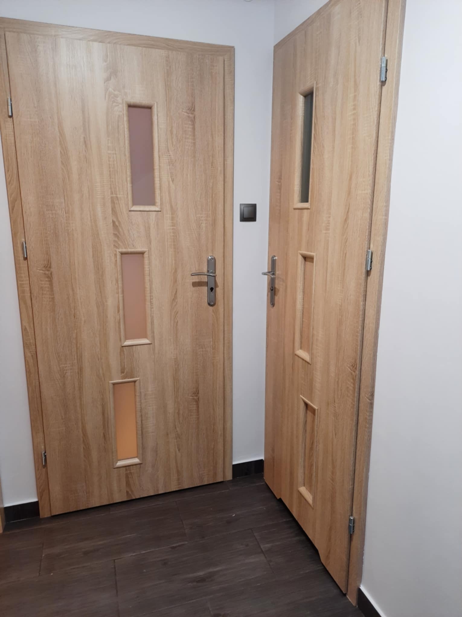Dwie drewniane drzwi wewnętrzne z podłużnymi przeszkleniami, widoczne klamki i włącznik światła na ścianie, ciemna podłoga.