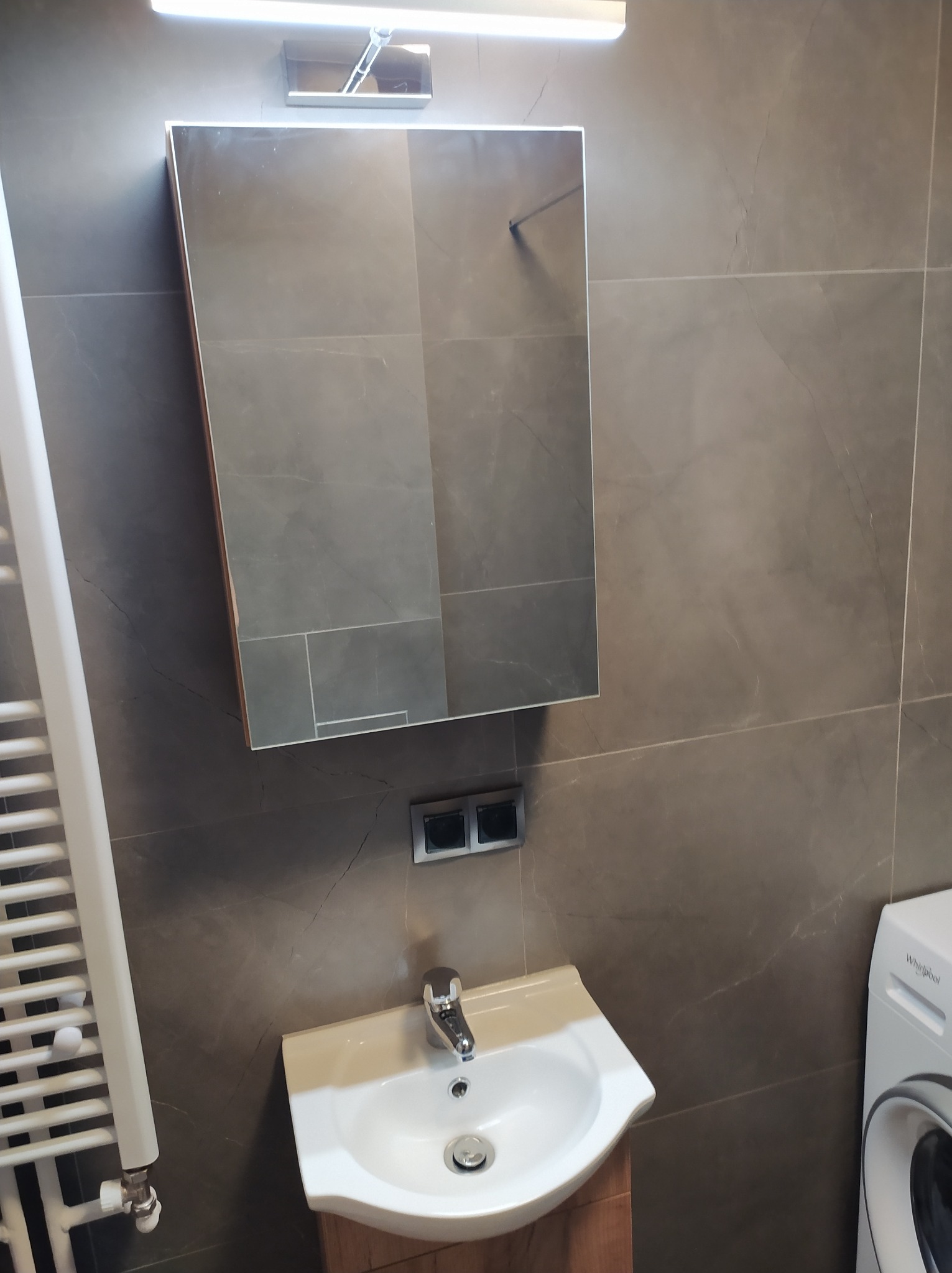 Nowoczesna łazienka z szarymi płytkami imitującymi marmur, lustro z oświetleniem LED, umywalka nablatowa z szafką z drewnopodobnego materiału, grzejnik drabinkowy i fragment pralki.