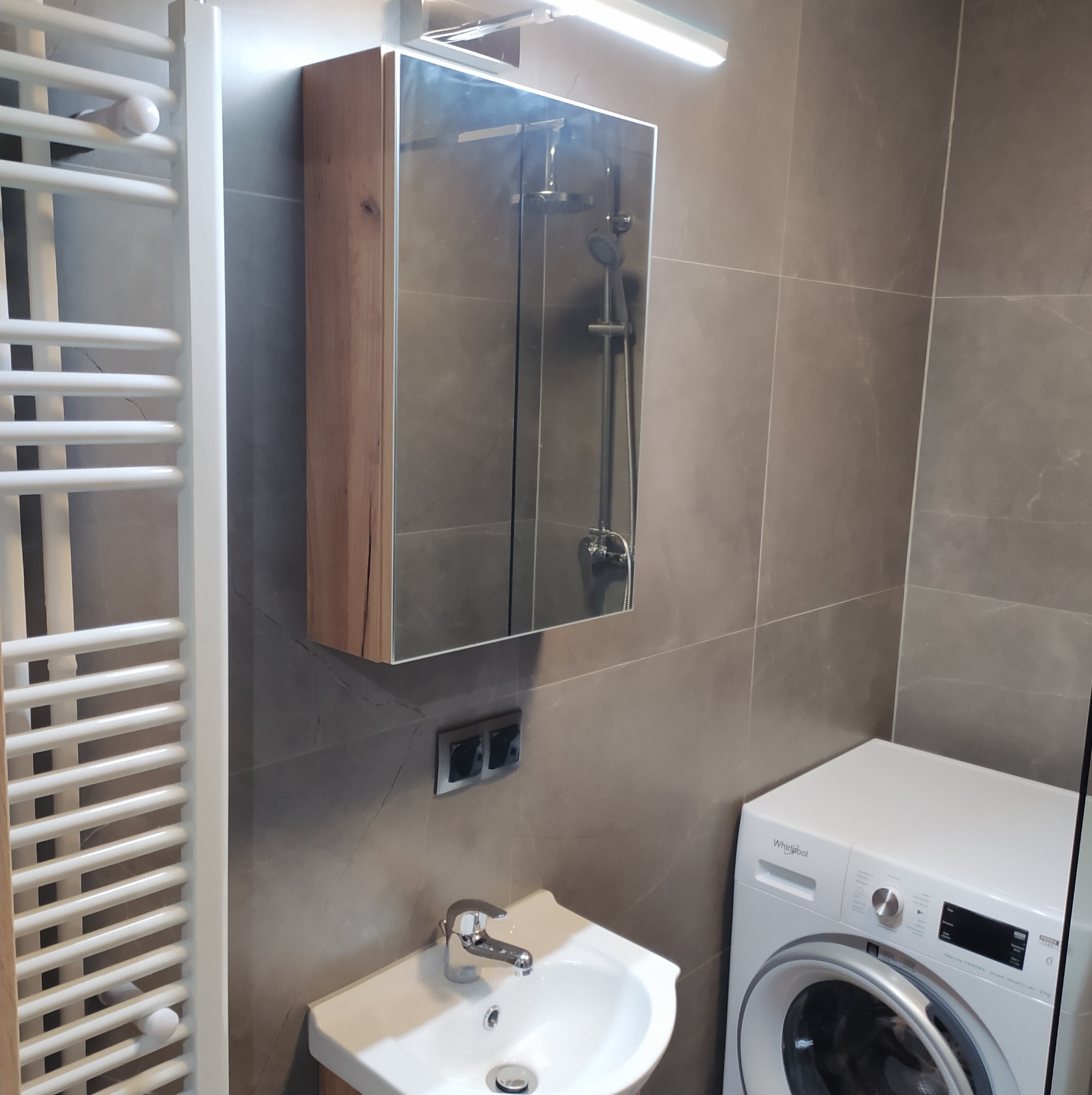 Nowoczesna łazienka z szarymi płytkami, grzejnikiem drabinkowym, umywalką, szafką z lustrem i pralką Whirlpool.