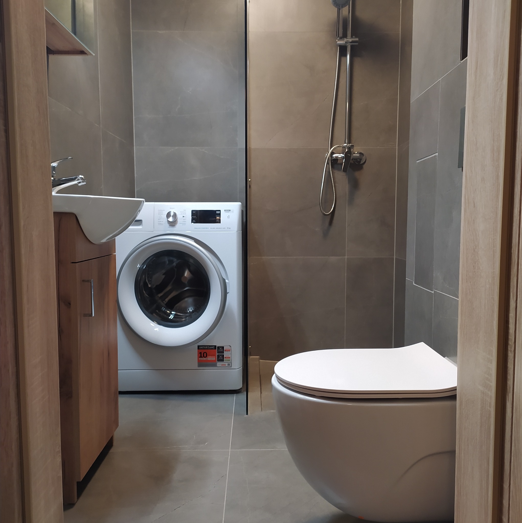 Nowoczesna łazienka z szarymi płytkami, wiszącą miską WC, szafką z umywalką z drewnopodobnym frontem i białą pralką automatyczną.
